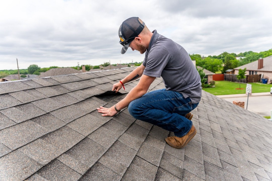 Roofer kneeling on an asphalt shingle roof to inspect lifted shingles