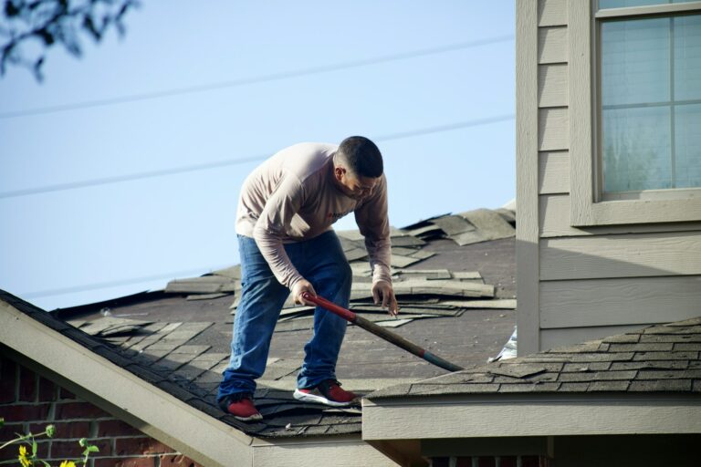Roofer using a pry bar to remove worn asphalt shingles from a house roof Roofer using a pry bar to remove worn asphalt shingles from a house roof