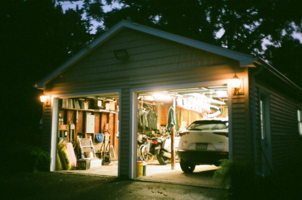 Detached garage with open doors and lights on at dusk, under a gable roof Detached garage with open doors and lights on at dusk, under a gable roof